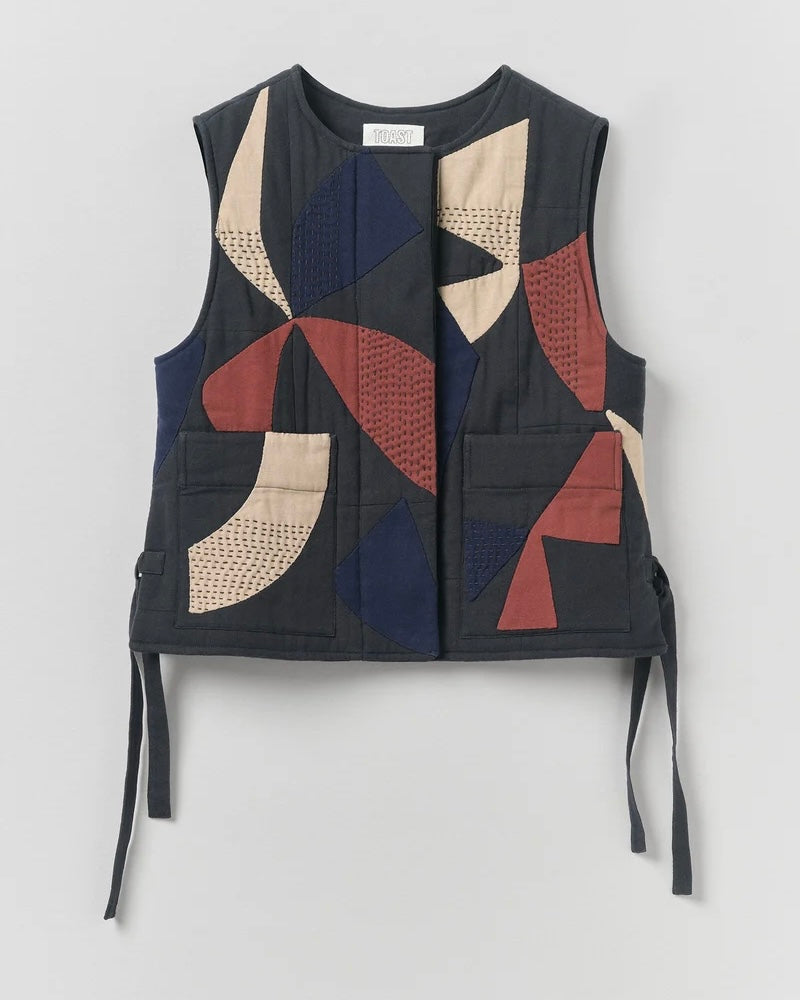 A sleeveless quilted vest with a geometric patchwork pattern in navy blue, terracotta red, beige, and black, featuring front pockets and side ties, shown against a light gray background.