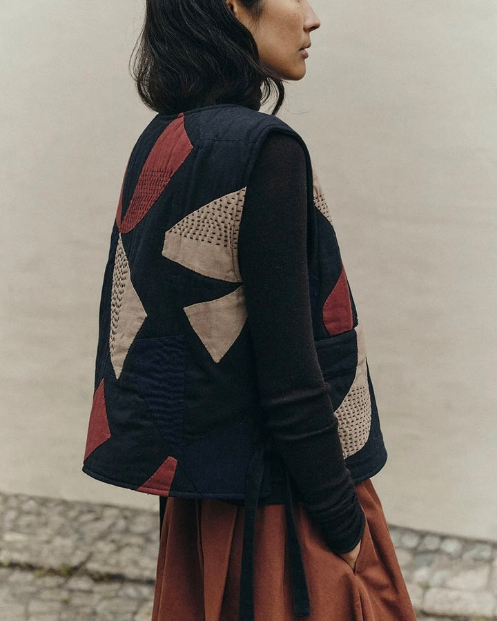 A woman shown from the back wearing a patchwork quilted vest over a black long-sleeve top and a rust-brown skirt, standing against a neutral beige wall.