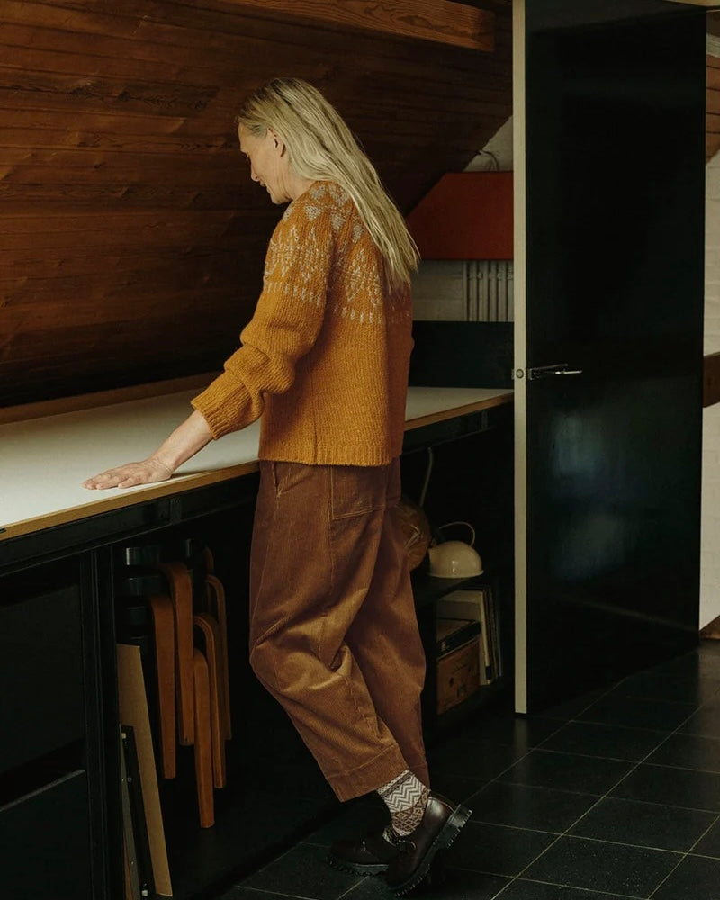 A woman stands in profile in a rustic room with wooden walls and a dark door, wearing a mustard yellow knit sweater with a white Nordic pattern, brown corduroy pants, and dark brown shoes.